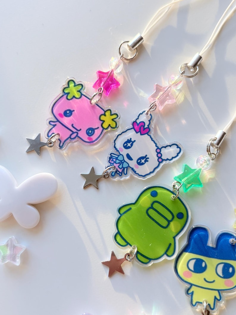 Op de afbeelding: Vier acryl telefoonhangers met kleurrijke stripfiguren, waaronder een roze wezen, een wit konijn, een groene blob en een blauw dier. Elke hanger is bevestigd aan een wit koord met kleine stervormige kralen en een zilverkleurige sterbedel.