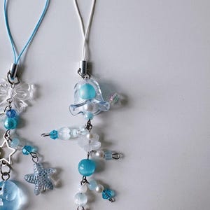 Puede incluir: Dos colgantes para teléfono con cuentas azules y blancas, abalorios transparentes y detalles de estrella. Un colgante tiene un cordón azul, el otro un cordón blanco. Los colgantes incluyen una estrella, una flor y un pequeño animal.