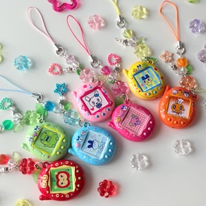 Puede incluir: Una colecci&oacute;n de siete llaveros de mascotas digitales de colores rojo, naranja, amarillo, verde claro, azul, rosa y magenta. Cada dispositivo tiene una pantalla con un personaje de dibujos animados y una correa de cuentas con dijes de flores.