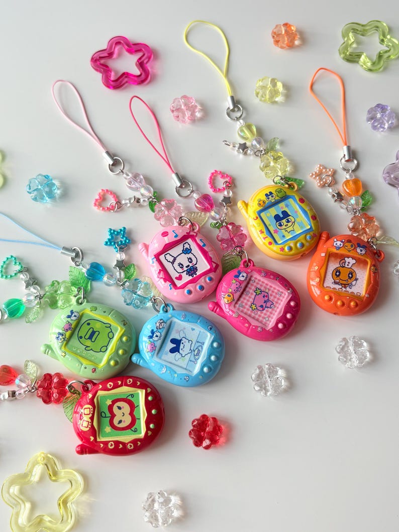 Puede incluir: Una colecci&oacute;n de siete llaveros de mascotas digitales de colores rojo, naranja, amarillo, verde claro, azul, rosa y magenta. Cada dispositivo tiene una pantalla con un personaje de dibujos animados y una correa de cuentas con dijes de flores.
