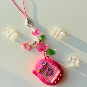 Puede incluir: Un llavero de mascota digital rosa brillante con una pantalla de patr&oacute;n de cuadros y un peque&ntilde;o personaje. El dispositivo est&aacute; unido a un cord&oacute;n rosa y a una cadena de dijes decorativos que consiste en cuentas de coraz&oacute;n rosa, cuentas claras y hojas verdes.