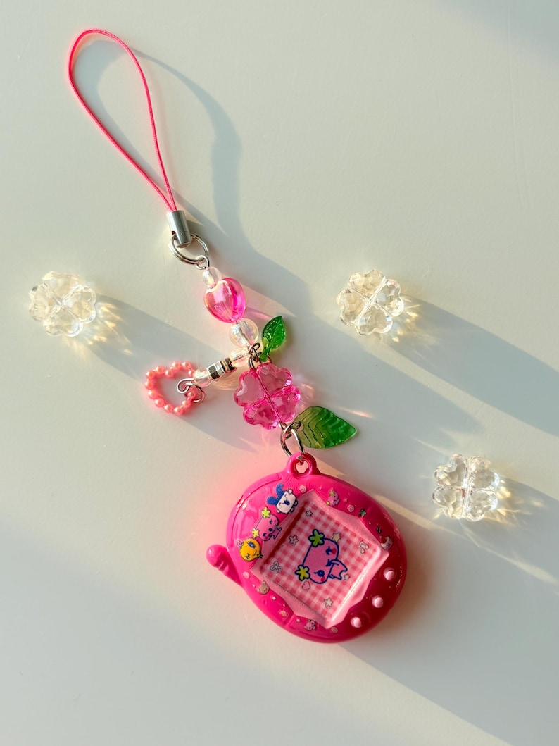 Puede incluir: Un llavero de mascota digital rosa brillante con una pantalla de patr&oacute;n de cuadros y un peque&ntilde;o personaje. El dispositivo est&aacute; unido a un cord&oacute;n rosa y a una cadena de dijes decorativos que consiste en cuentas de coraz&oacute;n rosa, cuentas claras y hojas verdes.
