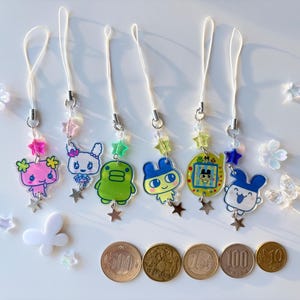 Op de afbeelding: Zes acryl telefoonhangers met kleurrijke stripfiguren, waaronder een roze wezen, een wit konijn, een groen figuur, een geel personage, een geel digitaal huisdierapparaat en een blauw konijn, elk met een wit koord en sterrenkralen.