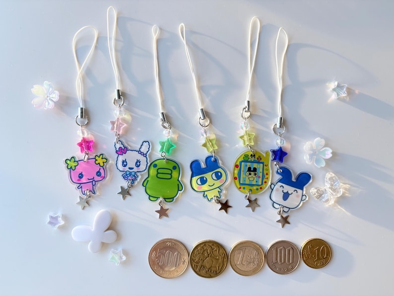 Op de afbeelding: Zes acryl telefoonhangers met kleurrijke stripfiguren, waaronder een roze wezen, een wit konijn, een groen figuur, een geel personage, een geel digitaal huisdierapparaat en een blauw konijn, elk met een wit koord en sterrenkralen.