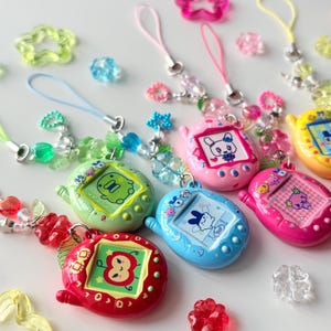 Puede incluir: Una colecci&oacute;n de llaveros de mascotas digitales coloridos en rojo, verde, azul, rosa y amarillo, cada uno con un personaje &uacute;nico en la pantalla. Los llaveros est&aacute;n unidos a cordones con cuentas y dijes en forma de estrella y coraz&oacute;n.