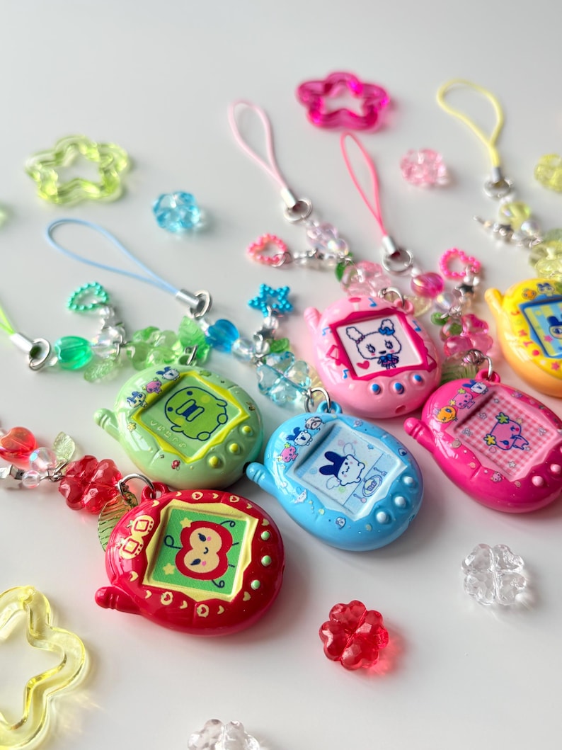 Puede incluir: Una colecci&oacute;n de llaveros de mascotas digitales coloridos en rojo, verde, azul, rosa y amarillo, cada uno con un personaje &uacute;nico en la pantalla. Los llaveros est&aacute;n unidos a cordones con cuentas y dijes en forma de estrella y coraz&oacute;n.