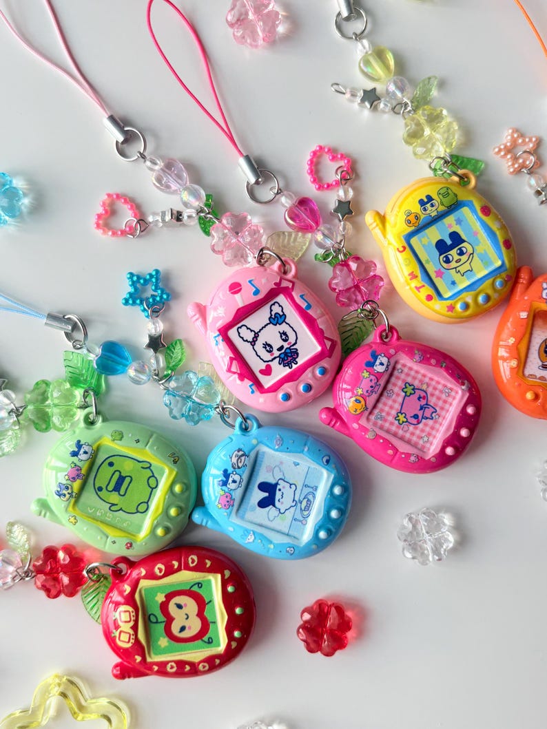 Puede incluir: Una colecci&oacute;n de llaveros de mascotas digitales coloridos con varias pantallas de personajes, incluyendo una manzana roja, un conejo azul y una criatura rosa. Cada dispositivo est&aacute; unido a un cord&oacute;n de cuentas con dijes de flores, corazones y estrellas.