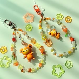 May include: Two beaded phone charms featuring orange bear figurines, clear and yellow flower-shaped beads, star-shaped beads, and translucent butterfly charms. Each charm includes a metal lobster clasp and a thin white cord for attachment.