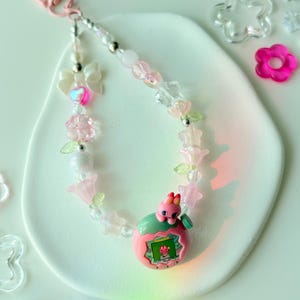 K&ouml;nnte beinhalten: Ein Schl&uuml;sselanh&auml;nger mit rosa und transparenten Perlen und einem rosafarbenen Verschluss. Er zeigt ein rosa-gr&uuml;nes elektronisches Spielzeug mit kleinem Bildschirm, umgeben von Perlen, Blumen und einer Schleife.