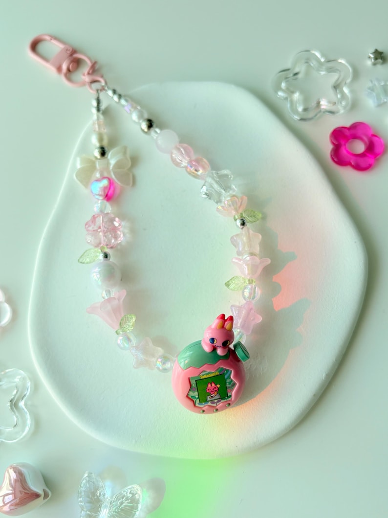 K&ouml;nnte beinhalten: Ein Schl&uuml;sselanh&auml;nger mit rosa und transparenten Perlen und einem rosafarbenen Verschluss. Er zeigt ein rosa-gr&uuml;nes elektronisches Spielzeug mit kleinem Bildschirm, umgeben von Perlen, Blumen und einer Schleife.
