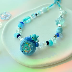 K&ouml;nnte beinhalten: Ein blaues Perlen-Schl&uuml;sselband mit einem kleinen, hellblauen elektronischen Spielzeug mit einem Bildschirm und einer kleinen blauen Kreatur oben. Das Schl&uuml;sselband enth&auml;lt verschiedene blaue und transparente Perlen, darunter Stern- und Blumenformen.
