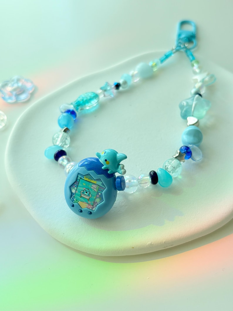 K&ouml;nnte beinhalten: Ein blaues Perlen-Schl&uuml;sselband mit einem kleinen, hellblauen elektronischen Spielzeug mit einem Bildschirm und einer kleinen blauen Kreatur oben. Das Schl&uuml;sselband enth&auml;lt verschiedene blaue und transparente Perlen, darunter Stern- und Blumenformen.