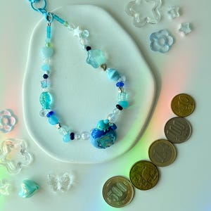 K&ouml;nnte beinhalten: Ein blaues Schl&uuml;sselband mit Perlen in verschiedenen Formen, darunter Sterne und Herzen. Das Schl&uuml;sselband hat einen blauen Anh&auml;nger und wird mit transparenten Stern- und Blumendekorationen sowie japanischen M&uuml;nzen pr&auml;sentiert. Die Perlen sind in Blaut&ouml;nen gehalten.