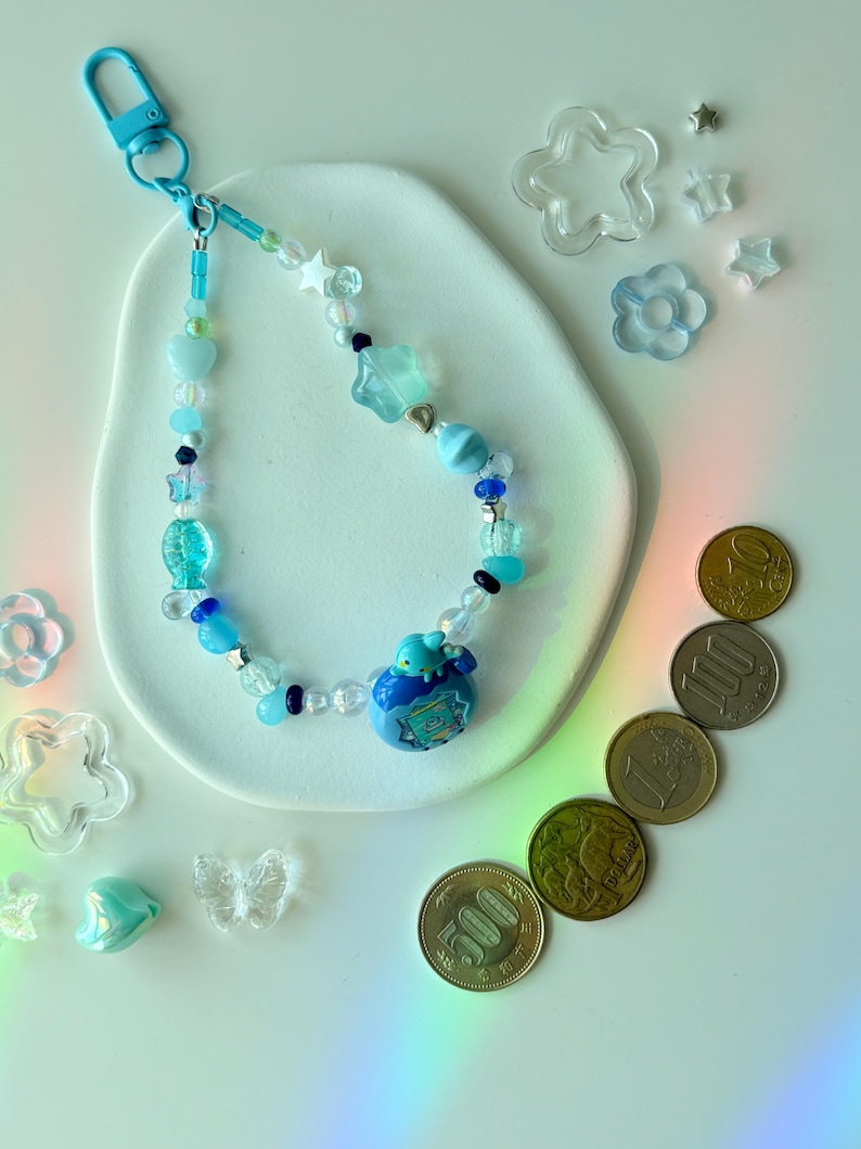 K&ouml;nnte beinhalten: Ein blaues Schl&uuml;sselband mit Perlen in verschiedenen Formen, darunter Sterne und Herzen. Das Schl&uuml;sselband hat einen blauen Anh&auml;nger und wird mit transparenten Stern- und Blumendekorationen sowie japanischen M&uuml;nzen pr&auml;sentiert. Die Perlen sind in Blaut&ouml;nen gehalten.