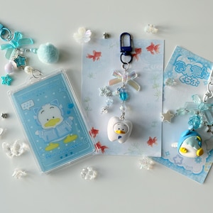 May include: A collection of whimsical keychains and a card holder. The keychains feature blue ribbons, star charms, and cartoon character pendants. The card holder has a cartoon duck design. The items are arranged on a white surface with decorative elements.
