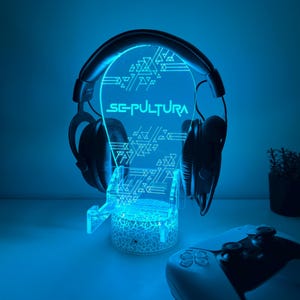 May include: A glowing blue headphone stand with the word "SEPULTURA" and geometric designs. Black headphones rest on the stand, and a white game controller is in the foreground. The stand is illuminated with blue light.