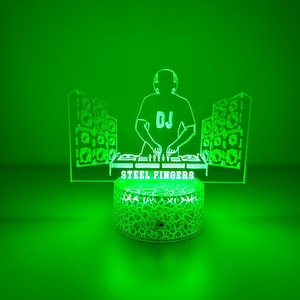 May include: A green LED lamp featuring a DJ silhouette with headphones, a DJ table, and speakers. The base of the lamp has a cracked glass design, and the text "STEEL FINGERS" is visible. The background is a solid green.