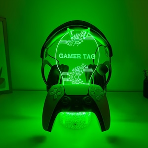 Könnte beinhalten: Ein grün beleuchtetes Gaming-Zubehör mit einem Controller- und Headset-Ständer. Der Ständer trägt den Text "GAMER TAG" in Weiß. Der Controller ist schwarz und weiß, und das Headset ist schwarz.