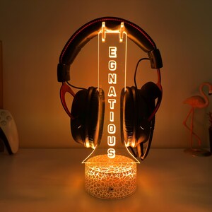 May include: A lighted headphone stand with the word "EGNATIOUS" vertically displayed. The stand holds a pair of black and red headphones. The base of the stand has a cracked glass design, and the overall color scheme is warm.