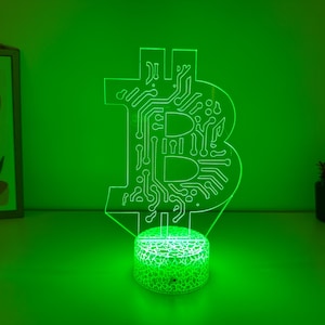 Pode incluir: Uma lâmpada LED verde em forma de símbolo Bitcoin. A lâmpada apresenta um design de placa de circuito dentro do símbolo Bitcoin e assenta numa base redonda e texturizada. O fundo é verde liso.