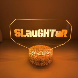 May include: A glowing LED lamp with the word "SLAUGHTER" in bold, orange letters. The lamp has a clear, geometric top and a textured, round base. The light emits a warm, orange glow, creating a striking visual effect.