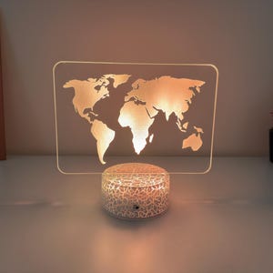 Puede incluir: Lámpara de noche con mapa del mundo iluminado. El mapa está grabado en un panel acrílico transparente, iluminado por una luz cálida. La base es una luz blanca, redonda y texturizada. La lámpara está sobre una superficie blanca.