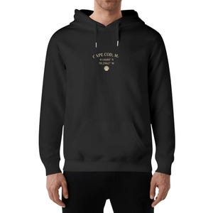 Op de afbeelding: Zwarte hoodie met de tekst "CAPE COD, M." en coördinaten 41.6688° N 70.2962° W, plus een schelpgrafiek. De hoodie heeft een voorzak en een capuchon met trekkoord.