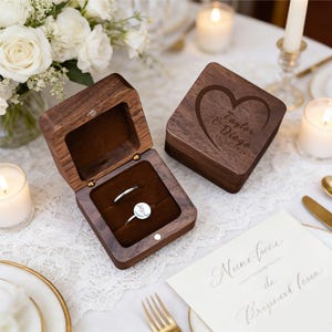 May include: Two wooden ring boxes, one open and one closed, sit on a white lace tablecloth. The open box reveals two rings nestled in a brown velvet interior. The closed box has a heart design with the names "Taylor & Diego" and a date engraved.