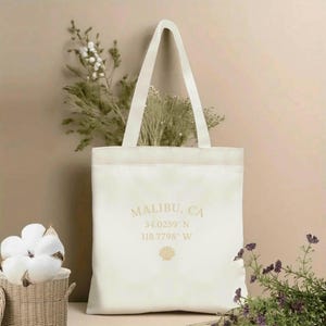 May include: Cream-colored tote bag with the text "MALIBU, CA" and coordinates, plus a seashell graphic. The bag has a rectangular shape with a wide opening and two long handles. The bag is made of a sturdy fabric and is suitable for carrying various items.