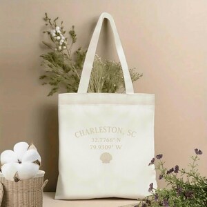 May include: Cream-colored tote bag with the text "Charleston, SC" and coordinates. The bag features a seashell graphic and has a rectangular shape with a handle. The bag is filled with greenery and is set against a neutral background.