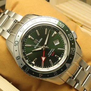 Könnte beinhalten: Eine Grand Seiko-Uhr aus Edelstahl mit grünem Zifferblatt und silbernem Armband. Die Uhr hat eine schwarz-grüne Lünette und wird auf einem gelben Kissen präsentiert. Das Zifferblatt zeigt den Text "Grand Seiko" und "GMT".
