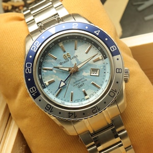 Puede incluir: Un reloj Grand Seiko GMT de acero inoxidable con esfera azul claro y bisel azul y plateado. El reloj tiene una pantalla de fecha y una pulsera de acero inoxidable. El reloj se muestra sobre una superficie amarilla.