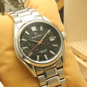 May include: A stainless steel Grand Seiko wristwatch with a black textured dial, silver hands, and a date display. The watch has a silver bracelet and is on a yellow cushion. The watch face reads "HI-BEAT 36000 80 HOURS". The watch is a luxury item.