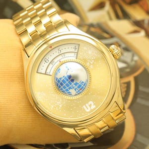 May include: Gold-toned wristwatch with a unique face design. The watch features a globe graphic at the center and the text "U2" at the bottom. The watch band is also gold-toned.