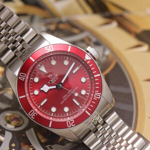 May include: A stainless steel wristwatch with a red dial and a matching red rotating bezel featuring white numerals. The watch has a silver-tone link bracelet, luminous hour markers, and the text Tudor Geneve 200m 660ft Master Chronometer.