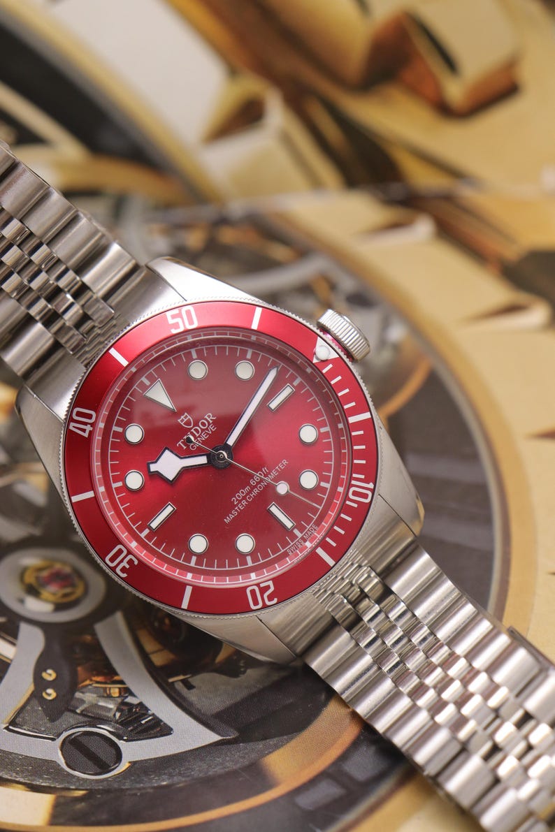 May include: A stainless steel wristwatch with a red dial and a matching red rotating bezel featuring white numerals. The watch has a silver-tone link bracelet, luminous hour markers, and the text Tudor Geneve 200m 660ft Master Chronometer.