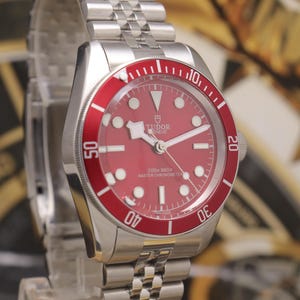 May include: A stainless steel wristwatch with a red dial and a matching red rotating bezel featuring white numerals. The watch has a silver-tone link bracelet, luminous hour markers, and the text Tudor Geneve 200m 660ft Master Chronometer.