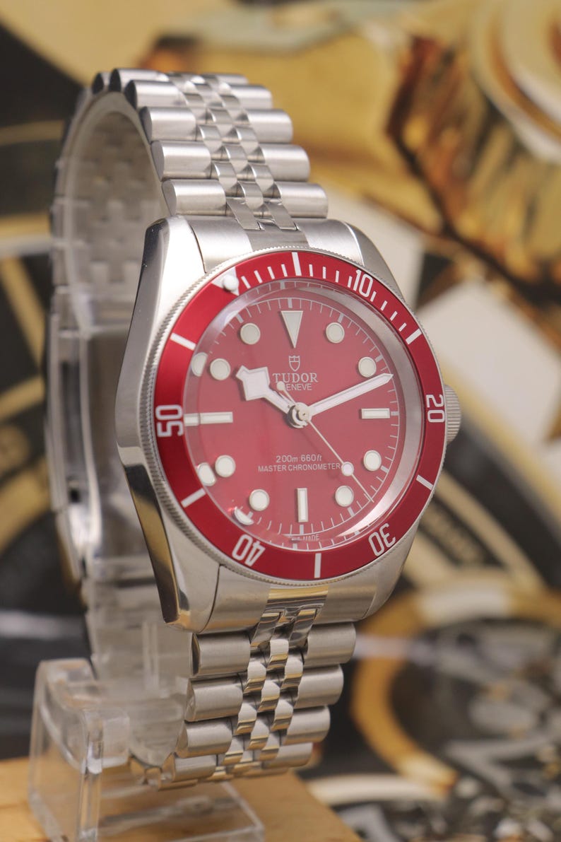 May include: A stainless steel wristwatch with a red dial and a matching red rotating bezel featuring white numerals. The watch has a silver-tone link bracelet, luminous hour markers, and the text Tudor Geneve 200m 660ft Master Chronometer.