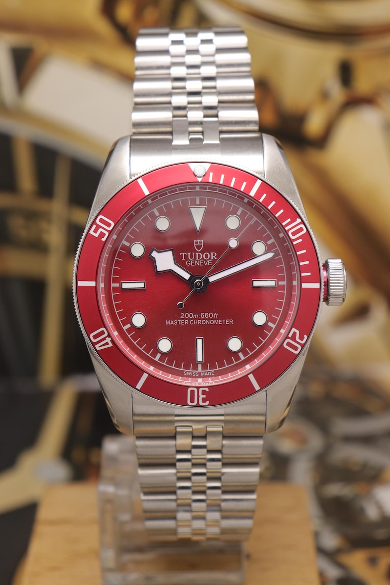 May include: A stainless steel wristwatch with a red dial and a matching red rotating bezel. The watch features a silver-tone link bracelet, luminous hour markers, and the text Tudor Geneve 200m 660ft Master Chronometer printed on the dial face.