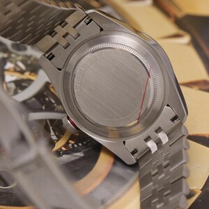 May include: A stainless steel wristwatch with a brushed metal case back and a multi-link bracelet. The case back features circular text engravings and a thin red line. A small red accent is visible on the side of the winding crown.