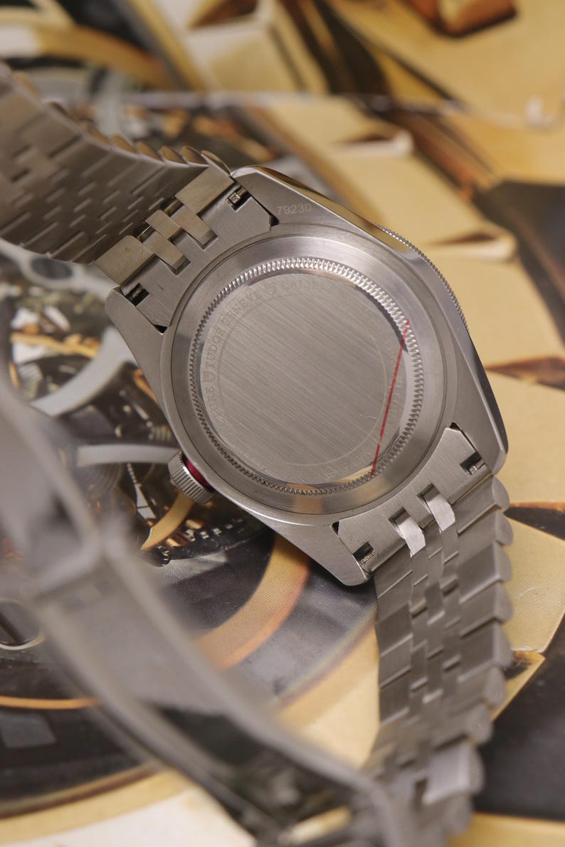 May include: A stainless steel wristwatch with a brushed metal case back and a multi-link bracelet. The case back features circular text engravings and a thin red line. A small red accent is visible on the side of the winding crown.
