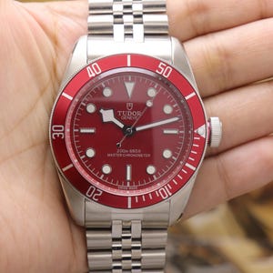 May include: A stainless steel wristwatch with a red dial and a matching red rotating bezel featuring white numerals. The watch has a silver-tone link bracelet, luminous hour markers, and the text Tudor Geneve 200m 660ft Master Chronometer.