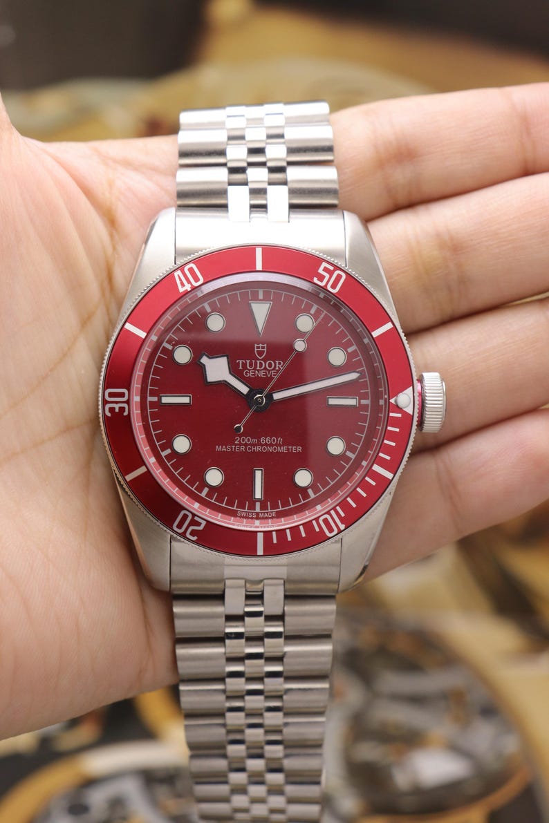 May include: A stainless steel wristwatch with a red dial and a matching red rotating bezel featuring white numerals. The watch has a silver-tone link bracelet, luminous hour markers, and the text Tudor Geneve 200m 660ft Master Chronometer.