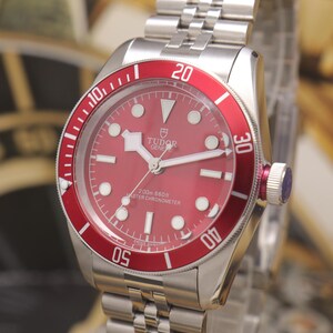 May include: A stainless steel Tudor wristwatch featuring a vibrant red dial and a matching red rotating bezel with white numerals. The watch includes a silver-tone link bracelet, luminous hour markers, and the text Tudor Geneve 200m 660ft Master Chronometer.