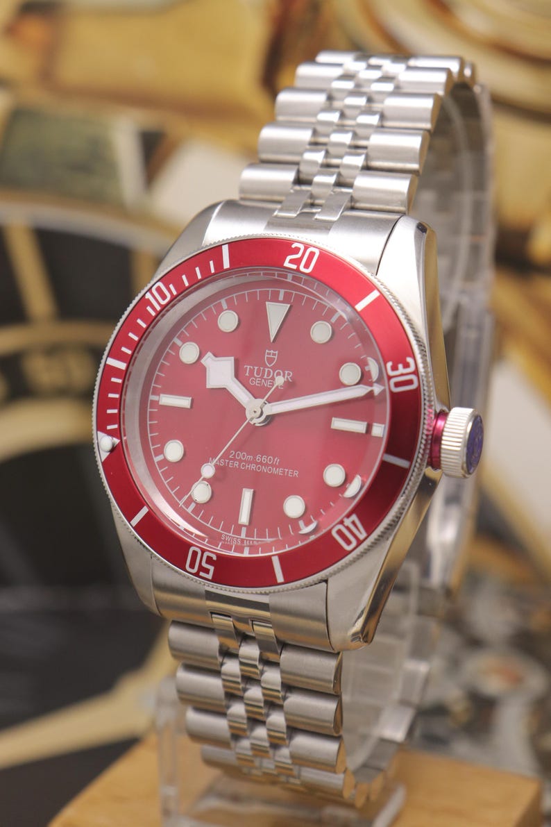 May include: A stainless steel Tudor wristwatch featuring a vibrant red dial and a matching red rotating bezel with white numerals. The watch includes a silver-tone link bracelet, luminous hour markers, and the text Tudor Geneve 200m 660ft Master Chronometer.