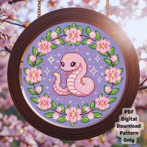 May include: A cross-stitch pattern featuring a pink snake with a crown, surrounded by pink flowers and green leaves, set against a lavender background. The design is framed in a round, dark wood frame. Text in the lower right corner reads "PDF Digital Download Pattern Only."