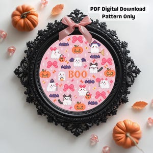May include: A framed cross-stitch pattern with a Halloween theme. The pink background features ghosts, cats, pumpkins, and bows. The word "BOO" is in the centre. The black frame is ornate, and the image is surrounded by pumpkins and decorative elements.