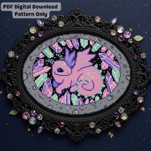 May include: A cross-stitch pattern featuring a pink dragon surrounded by crystals in shades of purple, green, and blue. The design is set within a black oval frame adorned with rhinestones. Text at the top reads "PDF Digital Download Pattern Only."