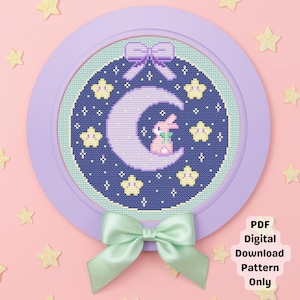 Può includere: Un motivo a punto croce con una luna a mezzaluna con un coniglietto rosa, circondato da stelle gialle su sfondo blu scuro. Il disegno è incorniciato in un cerchio lilla con un fiocco abbinato in alto e un fiocco verde menta in basso. Il testo recita "PDF Digital Download Pattern Only."