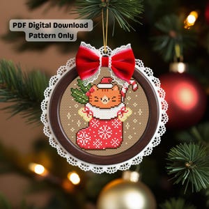 May include: A Christmas ornament featuring a cross-stitch design of a cat wearing a Santa hat and holding a candy cane. The ornament is in a wooden frame with a white lace edge and a red velvet bow. Text reads "PDF Digital Download Pattern Only."